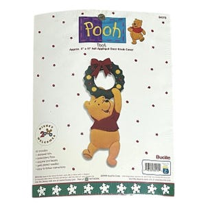 May include: A felt appliqué door knob cover featuring Winnie the Pooh holding a Christmas wreath. The package reads "Pooh" and "Approx. 4" x 11" Felt Appliqué Door Knob Cover". The kit includes stamped felts, embroidery floss, sequins, and beads.