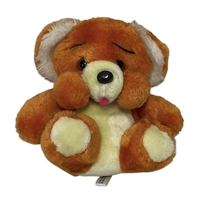 May include: A small, orange and cream plush teddy bear with a pink tongue sticking out. The bear has a sad expression and is sitting with its paws tucked in.