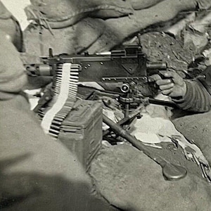 Pode incluir: Uma metralhadora preta com uma longa cinta de munição e um estoque de madeira. A arma está montada em um tripé e está sendo operada por uma pessoa.