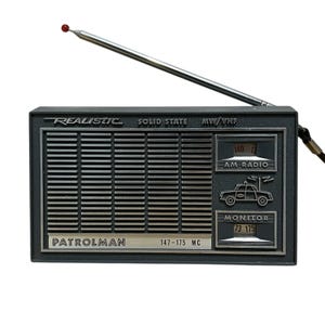 May include: A vintage Realistic Patrolman radio with a telescoping antenna. The dark gray radio features the text "Realistic", "Solid State", "MW/VHF", "AM Radio", and "Monitor". The radio has a silver speaker grill and a red-tipped antenna.