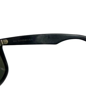 K&ouml;nnte beinhalten: Nahaufnahme einer schwarzen Sonnenbrille mit dem Text "B&L RAY-BAN U.S.A." auf dem B&uuml;gel. Die Sonnenbrille hat ein klassisches Design mit dunklem Rahmen und Gl&auml;sern. Die Scharnier- und B&uuml;geldetails sind sichtbar.