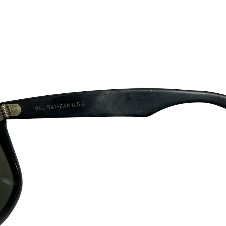 K&ouml;nnte beinhalten: Nahaufnahme einer schwarzen Sonnenbrille mit dem Text "B&L RAY-BAN U.S.A." auf dem B&uuml;gel. Die Sonnenbrille hat ein klassisches Design mit dunklem Rahmen und Gl&auml;sern. Die Scharnier- und B&uuml;geldetails sind sichtbar.