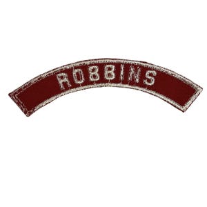 May include: A maroon, arched patch with the name "ROBBINS" embroidered in white thread. The patch has a silver border and is set against a white background. The shape is curved, and the text is centered.