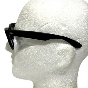 K&ouml;nnte beinhalten: Schwarze Sonnenbrille mit klassischem Design. Die Rahmen sind gl&auml;nzend schwarz und die Gl&auml;ser dunkel. Die Sonnenbrille wird auf einem wei&szlig;en Schaumstoffkopf pr&auml;sentiert, der das Seitenprofil der Brille zeigt.