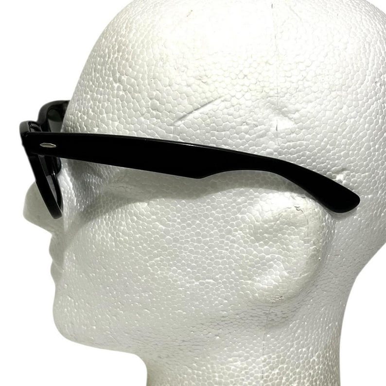 K&ouml;nnte beinhalten: Schwarze Sonnenbrille mit klassischem Design. Die Rahmen sind gl&auml;nzend schwarz und die Gl&auml;ser dunkel. Die Sonnenbrille wird auf einem wei&szlig;en Schaumstoffkopf pr&auml;sentiert, der das Seitenprofil der Brille zeigt.