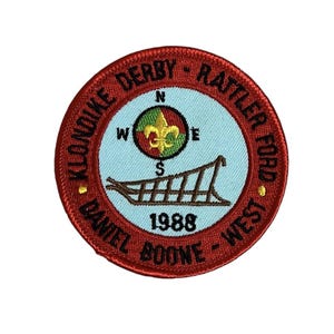 May include: An embroidered patch in red, white and blue with the text "Klondike Derby - Rattler Ford - Daniel Boone - West" and "1988". The patch features a compass with a fleur-de-lis and a sled.