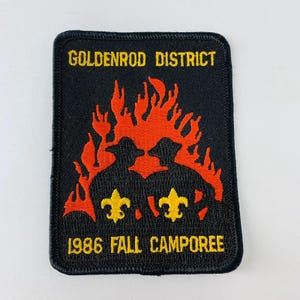 Może przedstawiać: Czarna prostokątna naszywka z żółtym napisem „GOLDENROD DISTRICT” i „1986 FALL CAMPOREE”. Naszywka przedstawia czerwony płomień nad dwoma żółtymi symbolami fleur-de-lis.