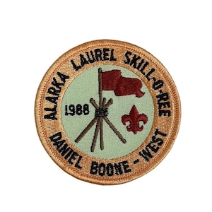 May include: A round, orange embroidered patch with the text "Alaska Laurel Skill-O-Ree Daniel Boone - West 1988" and a red flag on a stick with a scout symbol.