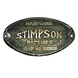 May include: An oval, antique-style metal sign with the text "MADE UNDER STIMPSON PATENTS SHOP NO. D880 276". The sign has a textured, dark gray background with gold-colored lettering and two holes for mounting.