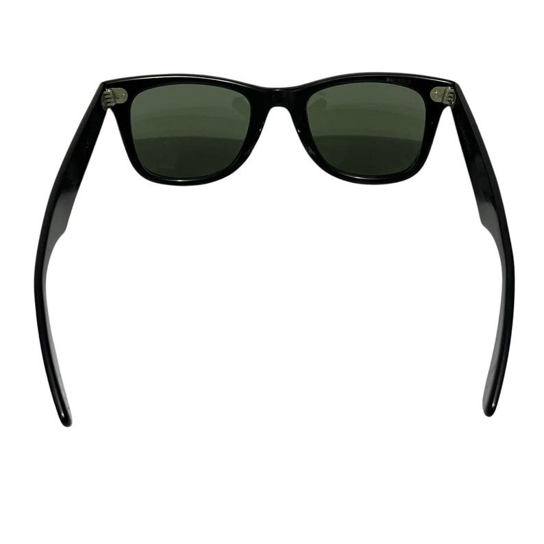 K&ouml;nnte beinhalten: Schwarze Sonnenbrille mit dunkelgr&uuml;nen Gl&auml;sern. Die Sonnenbrille hat ein klassisches rechteckiges Rahmendesign. Die B&uuml;gel der Sonnenbrille sind ausgeklappt und zeigen die Scharniere und die Form des Rahmens. Die Gl&auml;ser sind leicht reflektierend.
