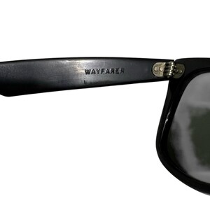 K&ouml;nnte beinhalten: Nahaufnahme einer schwarzen Wayfarer-Sonnenbrille. Die Sonnenbrille hat einen dicken, rechteckigen Rahmen und dunkle Gl&auml;ser. Das Wort "WAYFARER" ist auf dem linken B&uuml;gel eingepr&auml;gt. Das Scharnier ist sichtbar.