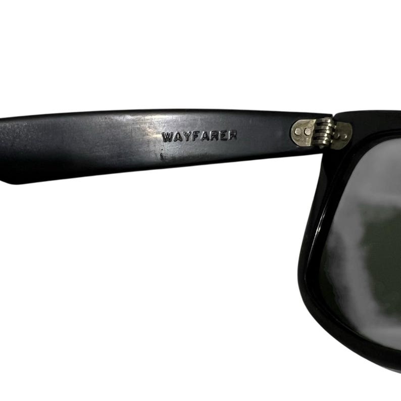 K&ouml;nnte beinhalten: Nahaufnahme einer schwarzen Wayfarer-Sonnenbrille. Die Sonnenbrille hat einen dicken, rechteckigen Rahmen und dunkle Gl&auml;ser. Das Wort "WAYFARER" ist auf dem linken B&uuml;gel eingepr&auml;gt. Das Scharnier ist sichtbar.