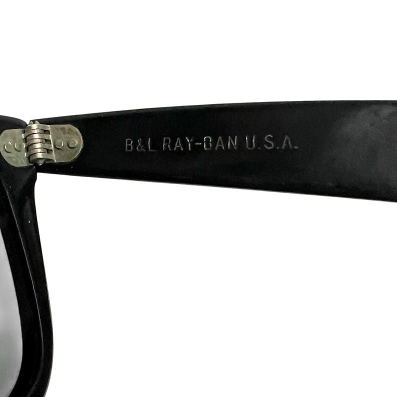 K&ouml;nnte beinhalten: Nahaufnahme eines schwarzen Sonnenbrillenb&uuml;gels mit dem wei&szlig;en Text "B&L RAY-BAN U.S.A.". Das Scharnier ist silberfarben. Die Sonnenbrille ist wahrscheinlich im Vintage- oder Retro-Stil.