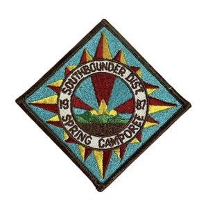 Może przedstawiać: Brązowa łata w kształcie rombu z niebieskim tłem i białym kołem w środku. Koło ma napis "1979 SOUTHBOUNDER DIST 1987" wokół zewnętrznej krawędzi i "SPRING CAMPOREE" wokół wewnętrznej krawędzi. Koło jest otoczone wzorem promieni słońca w kolorze czerwonym i żółtym.