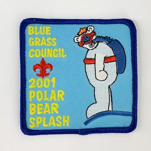 Puede incluir: Un parche cuadrado bordado con un borde azul y un fondo azul claro. El parche presenta un oso polar de dibujos animados con un esnórquel y una mochila. El texto en el parche dice "BLUE GRASS COUNCIL", "2001 POLAR BEAR SPLASH". También hay una flor de lis roja.