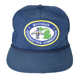 Puede incluir: Una gorra de béisbol azul con un parche bordado en amarillo, blanco y verde que dice "Michigan Aeronautics Commission" con una estrella a cada lado del texto y un pequeño avión en el centro.