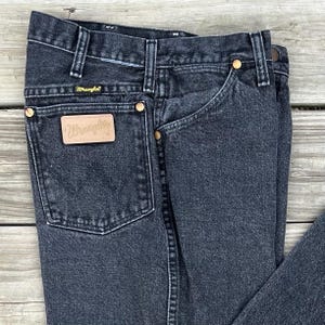 May include: Close-up of black denim Wrangler jeans. The jeans have a classic five-pocket design with a leather patch and gold-colored rivets. The Wrangler logo is visible on the back pocket and the leather patch.