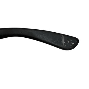 K&ouml;nnte beinhalten: Nahaufnahme eines schwarzen Brillenb&uuml;gels. Der B&uuml;gel ist gebogen und hat eine matte Oberfl&auml;che. Kleine, hellfarbige Unvollkommenheiten sind auf der Oberfl&auml;che sichtbar. Der Hintergrund ist schlicht wei&szlig;.