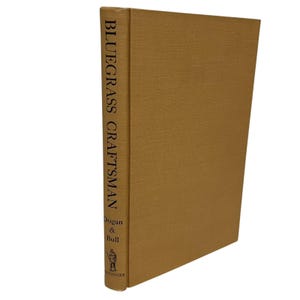 May include: A hardcover book titled "Bluegrass Craftsman" with a tan cover. The book's spine displays the title vertically, along with the authors' names, Dugan & Bull, and a small illustration. The book is set against a white background.