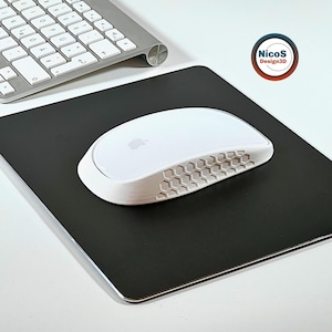 Könnte beinhalten: Eine weiße Computermaus mit glatter Oberfläche, Apple-Logo und einer strukturierten Seitenwand mit hexagonalem Wabenmuster. Die Maus liegt auf einem quadratischen schwarzen Mauspad neben einer silbernen kabellosen Tastatur.