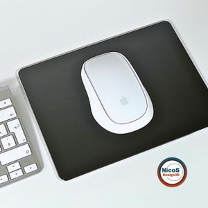 Könnte beinhalten: Ein rechteckiges schwarzes Mauspad mit abgerundeten Ecken, einer weißen Computermaus mit Apple-Logo auf der Oberseite und einem Teil einer weißen Tastatur auf der linken Seite. Ein kreisförmiges Logo mit dem Text NicoS Design3D ist zu sehen.