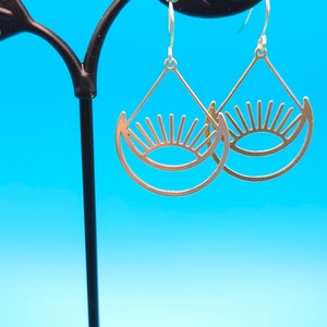 May include: A pair of gold-toned earrings with a sun and moon design. The earrings are hanging from a black earring stand against a blue background.