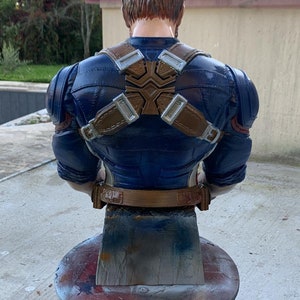 K&ouml;nnte beinhalten: Eine blaue und braune Captain America B&uuml;ste. Die B&uuml;ste ist mit verschr&auml;nkten Armen dargestellt und tr&auml;gt einen blauen und roten Anzug mit einem braunen G&uuml;rtel und Tr&auml;gern. Die B&uuml;ste steht auf einem runden, grauen Metallst&auml;nder.