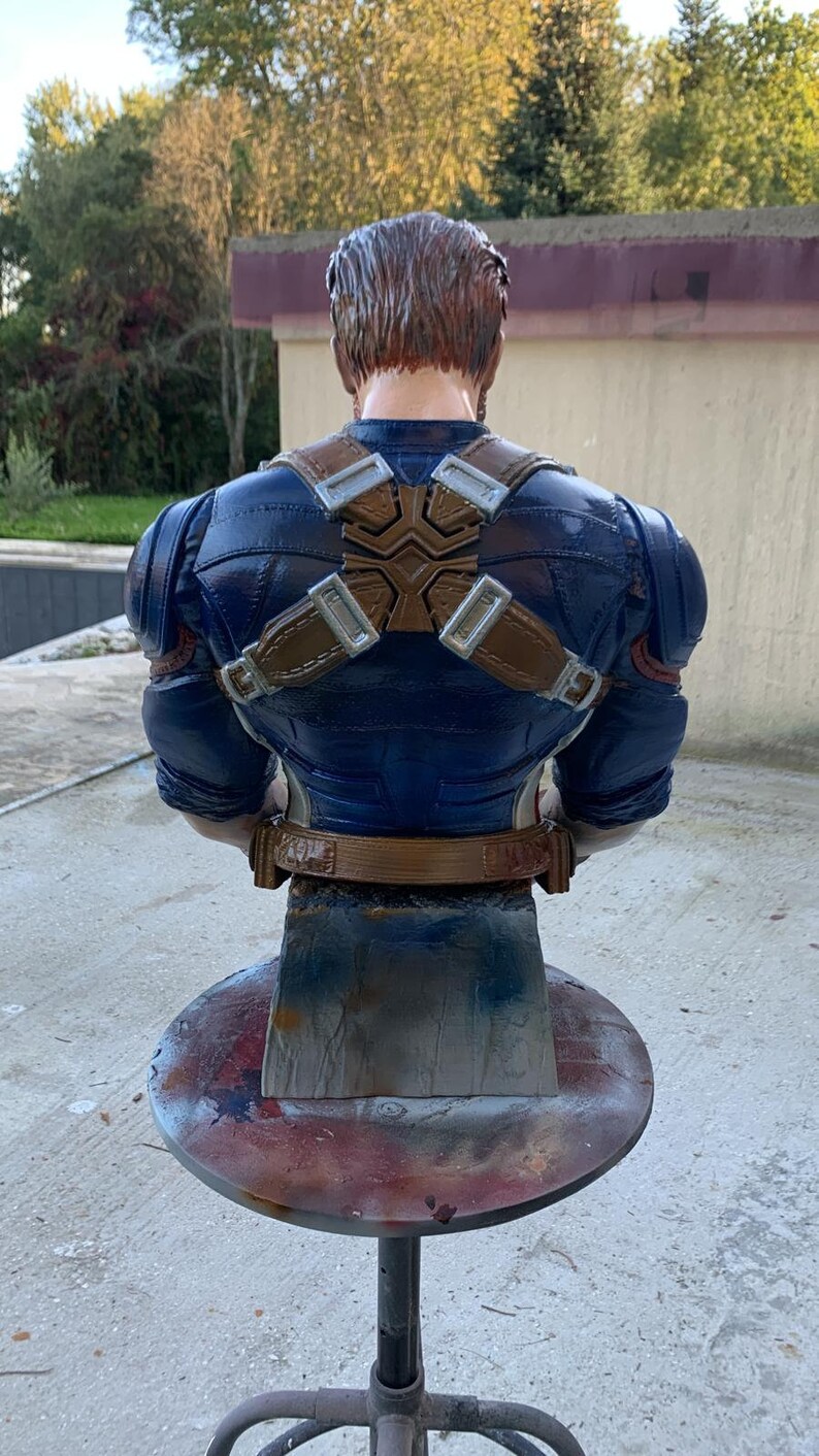 K&ouml;nnte beinhalten: Eine blaue und braune Captain America B&uuml;ste. Die B&uuml;ste ist mit verschr&auml;nkten Armen dargestellt und tr&auml;gt einen blauen und roten Anzug mit einem braunen G&uuml;rtel und Tr&auml;gern. Die B&uuml;ste steht auf einem runden, grauen Metallst&auml;nder.