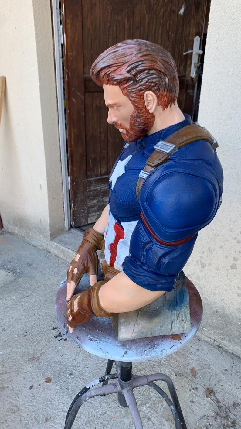 K&ouml;nnte beinhalten: Eine blau und rot bemalte B&uuml;ste von Captain America, die einen braunen Lederg&uuml;rtel und Handschuhe tr&auml;gt. Die B&uuml;ste sitzt auf einem grauen Hocker.