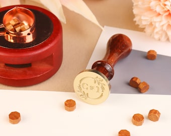 Könnte beinhalten: Ein goldfarbener Wachssiegel-Stempel mit einem floralen Design und den Initialen "C & J", die von einem Kranz umgeben sind. Der Stempel ist an einem Holzgriff befestigt. Auf einer weißen Oberfläche liegen außerdem mehrere braune Wachssiegel-Pellets.
