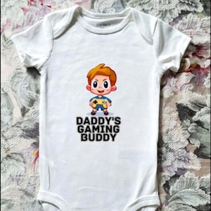May include: White baby onesie with a cartoon graphic of a child holding a game controller. The text "DADDY'S GAMING BUDDY" is printed below the graphic in black letters. The onesie has short sleeves and snap closures.
