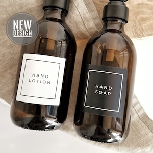 Op de afbeelding: Twee amberkleurige glazen flessen met zwarte pompdoseerders. Eén fles heeft een wit vierkant etiket met de tekst HAND LOTION, de andere een zwart vierkant etiket met de tekst HAND SOAP. Een ronde badge in de hoek vermeldt de tekst NEW DESIGN.