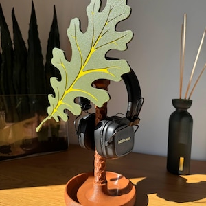 Op de afbeelding: Een bruine hoofdtelefoonstandaard met een bladvorm, met een zwarte hoofdtelefoon. Het blad is groen met gele accenten. De standaard heeft een ronde basis en een getextureerde steel. De hoofdtelefoon is voorzien van "Roseland".