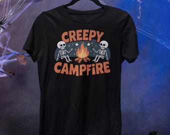 Może przedstawiać: Czarny t-shirt z grafiką o tematyce Halloween. Projekt przedstawia pomarańczowy napis "CREEPY CAMPFIRE" z ilustracjami szkieletów opiekaniem pianek przy ognisku. Koszulka wisi na czarnym wieszaku.