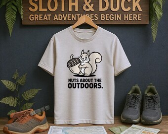 May include: A light beige t-shirt with a cartoon squirrel holding an acorn, with the text "NUTS ABOUT THE OUTDOORS." The shirt is displayed on a wooden hanger. Hiking shoes, a map, and a sign that says "SLOTH & DUCK GREAT ADVENTURES BEGIN HERE" are also visible.