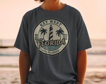 May include: Dark gray t-shirt featuring a circular graphic with the text "Key West Florida Established 1828." The design includes a lighthouse, palm trees, and ocean waves. The shirt is a casual wear item.