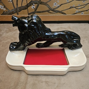Può includere: Figurina di leone in ceramica nera lucida, su una base rettangolare bianca con interno rosso. Il leone è in posizione reclinata, con la testa sollevata e la bocca aperta. La base ha un design curvo.