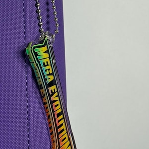 May include: A purple pencil case with a silver chain and a rectangular, iridescent charm that reads "MEGA EVOLUTION" in yellow and green. The charm is attached to the pencil case with a silver chain.