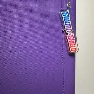 May include: Purple zippered binder with a clear plastic charm attached to it. The charm has the words "Sword & Shield" printed on it in a rainbow gradient.