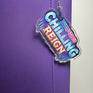 May include: A purple binder with a matching charm. The charm is clear with the words "Chilling Reign" in blue and orange, with the "Sword & Shield" logo. The binder has a textured surface and a silver chain.