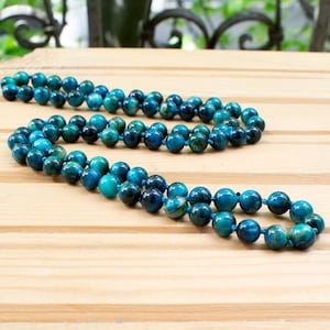 Puede incluir: Dos collares de cuentas con cuentas de color turquesa y azul oscuro. Las cuentas redondas están ensartadas juntas, sobre una superficie de madera marrón claro. El fondo está borroso, con un vistazo a una barandilla de metal verde y negro.