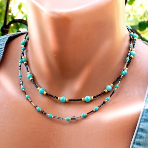 Può includere: Una collana a più fili con piccole perline nere, perline rotonde color turchese e distanziatori metallici color oro. Il design incorpora sottili perline cilindriche scure distribuite lungo i fili per un aspetto a strati.