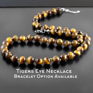 Puede incluir: Un collar hecho de cuentas de ojo de tigre pulidas, con una mezcla de tonos marrones y dorados. El collar está ensartado en un cordón y tiene un cierre plateado. El texto "Tigers Eye Necklace Bracelet Option Available" se muestra debajo.