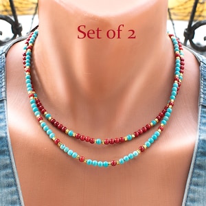 May include: A set of 2 necklaces featuring small round turquoise and deep red beads interspersed with tiny gold-toned spacers. The text Set of 2 is visible in red font above the necklaces, which are arranged in a layered style around the neck.