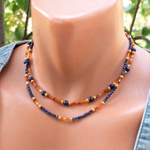Puede incluir: Un collar de doble hebra con pequeñas cuentas facetadas de color azul oscuro, cuentas naranjas translúcidas y cuentas espaciadoras de tono dorado metálico. El diseño alterna entre los segmentos azules y naranjas, creando un aspecto superpuesto.