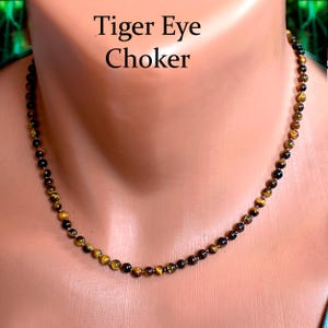 May include: A beaded choker necklace made of tiger eye stones. The necklace features small, round beads in shades of brown, gold, and black. The text "Tiger Eye Choker" is displayed above the necklace.