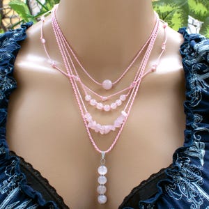 以下が含まれることがあります： さまざまなサイズのローズクォーツビーズが付いた、マルチストランドのピンクのネックレス。 ネックレスは重ね着されており、さまざまな長さのストランドがあります。 下のストランドには、ローズクォーツビーズの垂直配置があります。 ネックレスは濃い青色のトップスの上に着用されています。