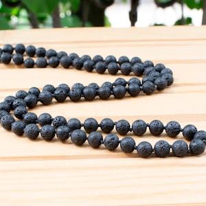 Puede incluir: Un collar largo hecho de cuentas de roca volcánica negra. Las cuentas son redondas y tienen una textura porosa. El collar se exhibe sobre una superficie de madera clara, resaltando las cuentas oscuras.
