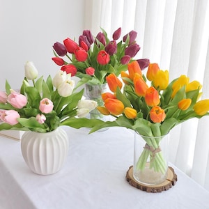May include: Arrangement of colorful tulips in vases. The flowers are in shades of pink, white, red, orange, yellow, and purple. The vases are clear glass and white ceramic. The arrangement is on a white surface.
