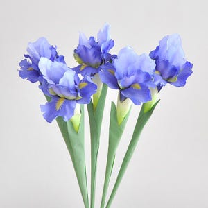 May include: A bouquet of artificial iris flowers. The flowers have layered, light blue petals with yellow accents at the center. The stems are green and the overall arrangement is set against a white background.